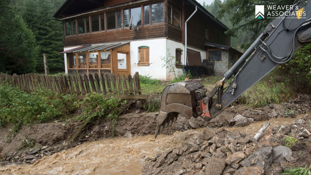 An excavator bucket clearing mud and debris from a fast-flowing mudslide next to a two-story home